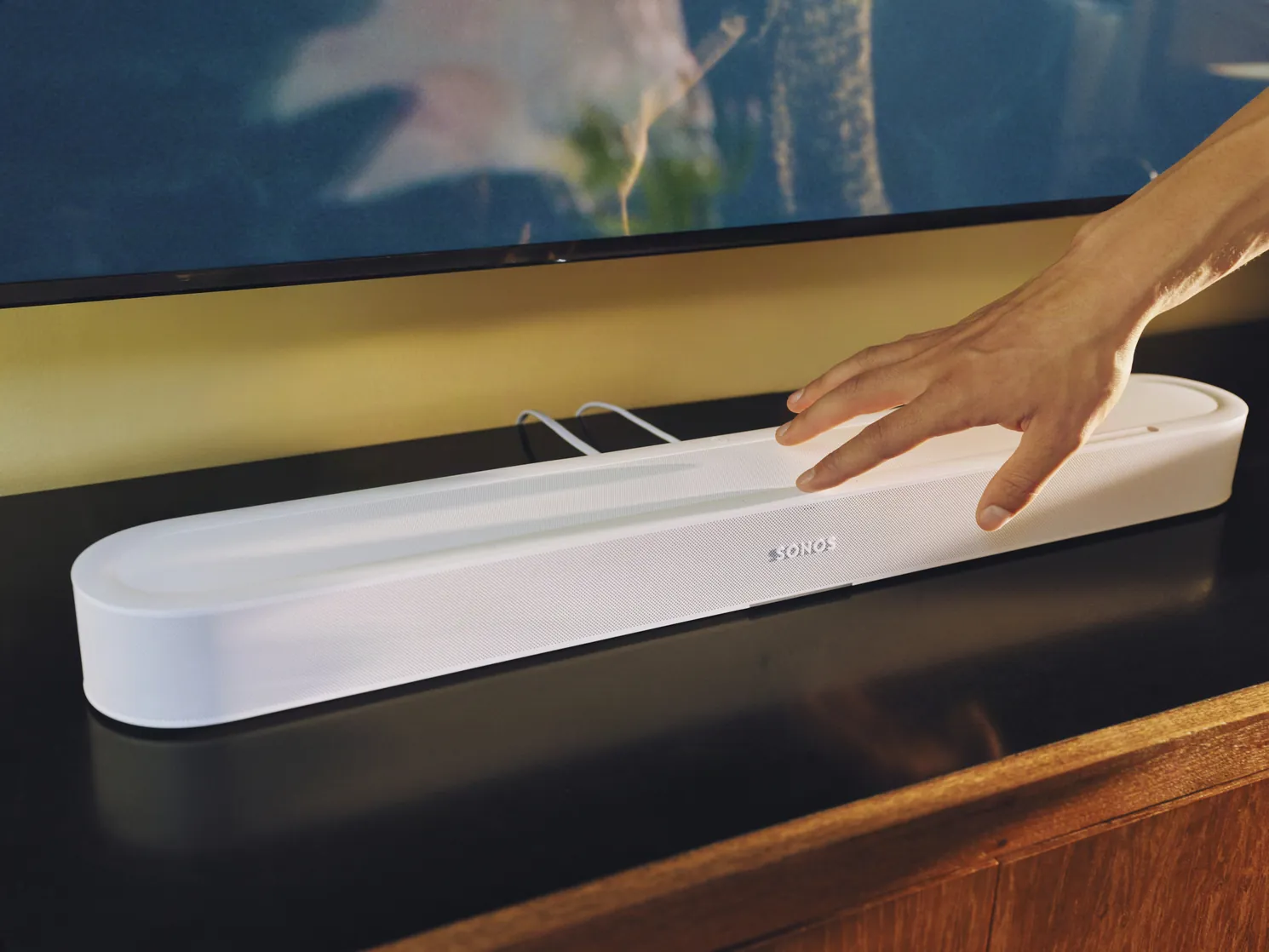 Hand op de aanraakbediening bovenop een witte Sonos Beam Ultra onder een tv