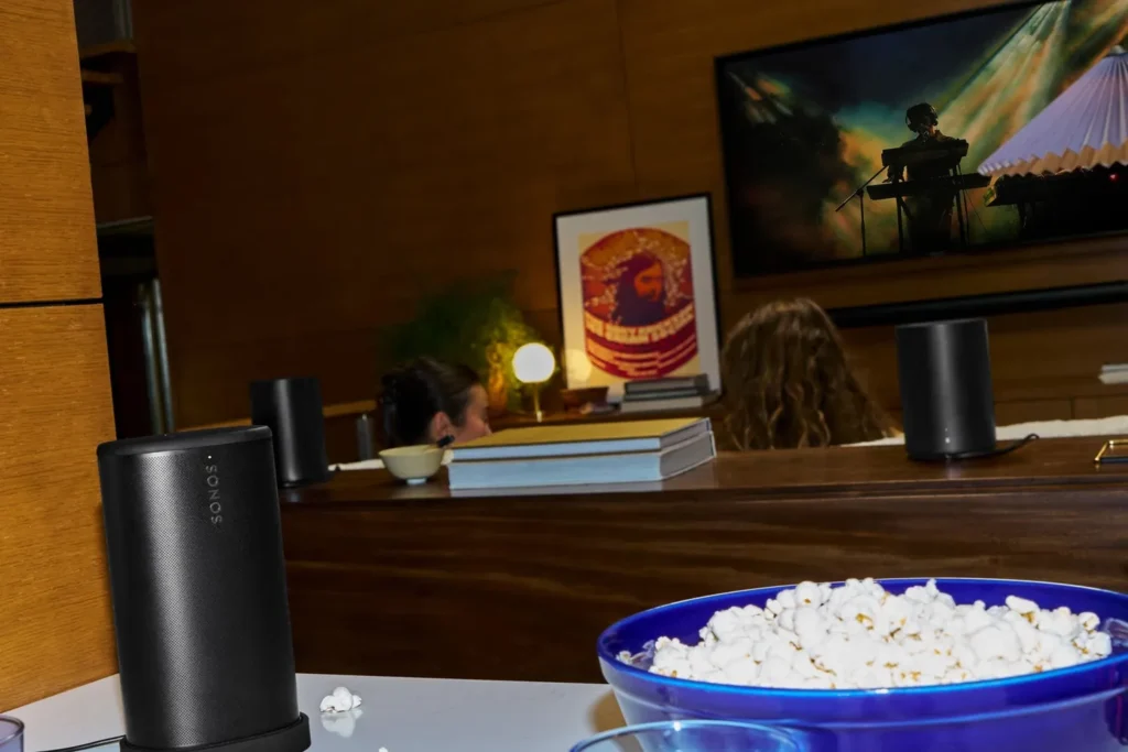 Zwarte Sonos Play op zijn oplaadstation naast een schaal popcorn, met de tv op de achtergrond